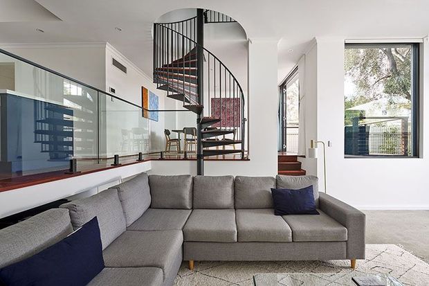 Bright contemporary living room with a large grey L-shaped sectional sofa, white walls and ceiling, and a striking black spiral staircase with timber treads. A glass balustrade separates the living area from an elevated open section, while large windows provide views of greenery outside. Colourful wall art and dark blue cushions add restrained accents.
