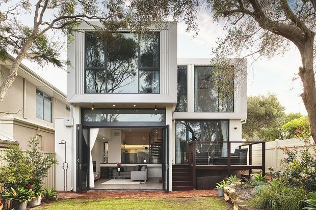 Exterior rear view of a contemporary multi-level residence with a crisp white facade, extensive dark-framed glazing, and a large open ground-floor living area. A timber deck with dark horizontal privacy screening projects from the right side, while mature trees and landscaped planting surround the home.