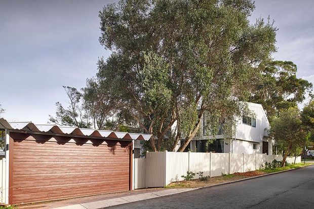 Street-facing exterior view of a contemporary multi-storey suburban residence. The home has white rendered walls, dark-framed windows, and a corrugated metal upper roof or wall element, partially screened by mature trees and a white vertical-panel fence. A separate timber-toned garage with a distinctive repeated triangular corrugated roofline fronts the street, alongside a paved footpath and asphalt road.