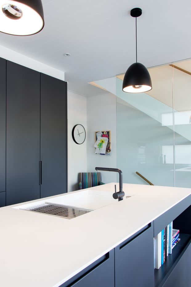 Bright contemporary kitchen with a large white island benchtop, integrated sink and dark matte cabinetry. Black pendant lights hang above the island, while a full-height glass balustrade reveals a staircase beyond. A compact dining or seating area and wall-mounted clock add colour and function.