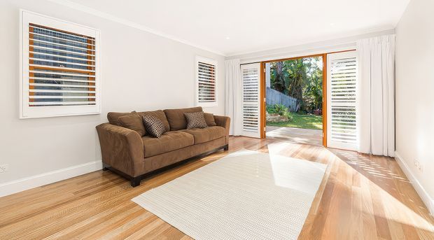 Bright, airy living room with a brown upholstered sofa, pale area rug, and polished light timber flooring. White walls and sheer curtains surround timber-framed louvred windows and glazed folding or French-style doors opening to a garden, creating strong natural light and indoor-outdoor connection.