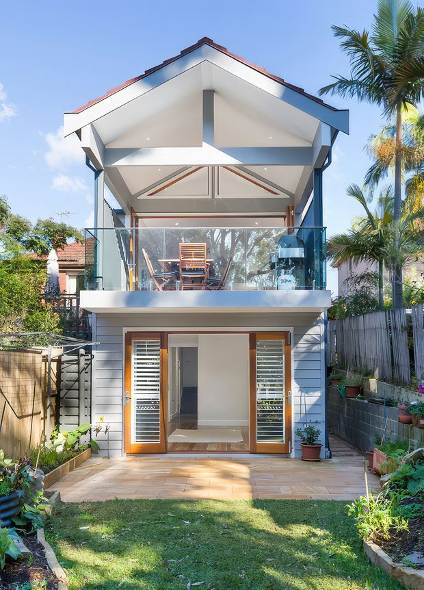 A bright two-storey residential rear facade viewed from the garden. The compact home has a steep gabled roof with exposed structural framing, white horizontal weatherboard cladding, timber-framed glazed doors, a projecting upper-level balcony with frameless glass balustrade, and a paved terrace opening onto a landscaped garden. Tropical palms and surrounding planting are visible around the property.