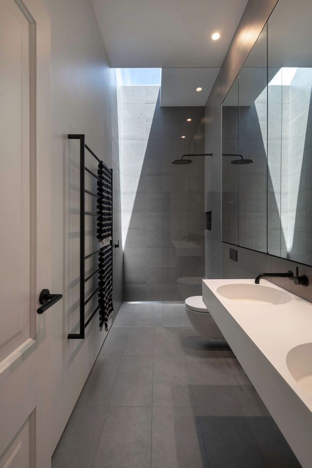 A narrow contemporary bathroom with a walk-in shower at the rear, wall-mounted rain shower fittings, a wall-hung toilet, a long white double vanity, and a large mirrored wall. Grey porcelain tile covers the floor and shower walls, while a skylight or clerestory opening brings in strong natural light. A black heated towel rail and matte-black tapware provide contrast against the pale finishes.