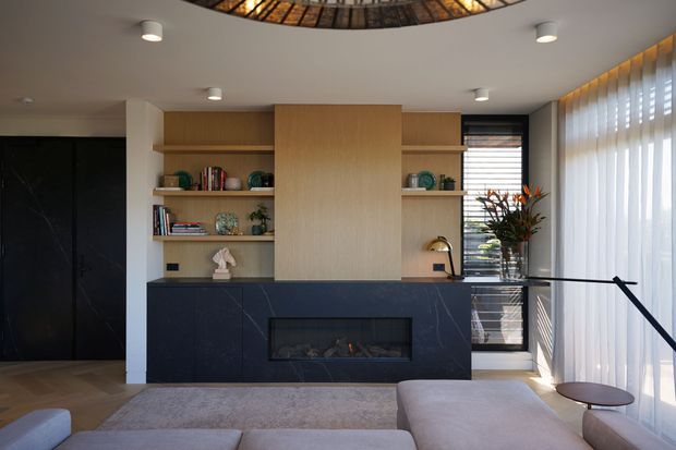 Contemporary living room featuring a full-height built-in entertainment wall with warm timber shelving and panelled cabinetry above dark stone-look cabinetry and a linear fireplace. A large sectional sofa, occasional table, sheer curtains, glazing, and a sculptural circular ceiling feature complete the refined interior.