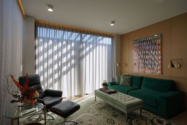 A refined living room with a deep green sofa, black leather lounge chair and ottoman, glass coffee tables, and a patterned upholstered bench. Full-height sheer curtains filter strong daylight across the room, while warm timber wall panelling, integrated lighting, brass-toned lamps, and colourful abstract artwork add a sophisticated layered finish.