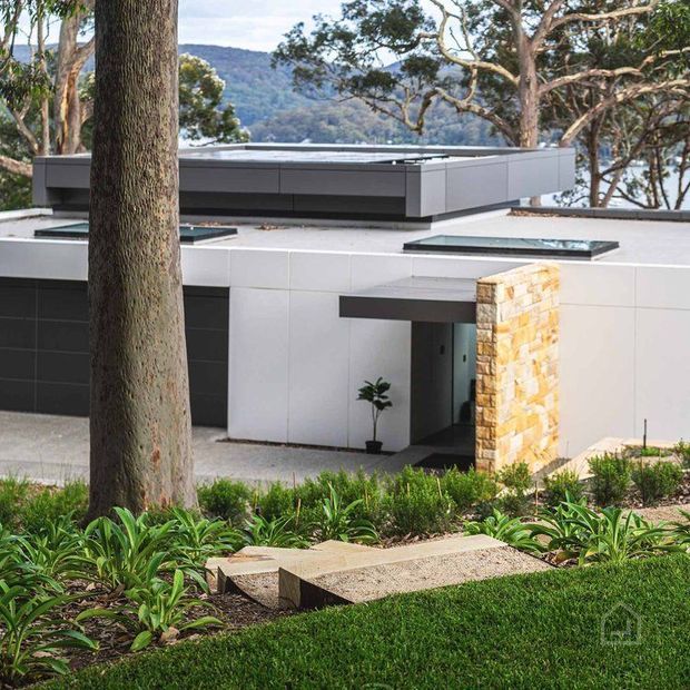 Exterior view of a contemporary flat-roofed house set into a landscaped, sloping site. The building features clean white panelled walls, dark charcoal horizontal cladding and roof elements, expansive low-profile glazing, and a prominent stacked-stone entry wall. Mature trees and planted garden beds frame the approach, with a mountain or bushland backdrop visible beyond.