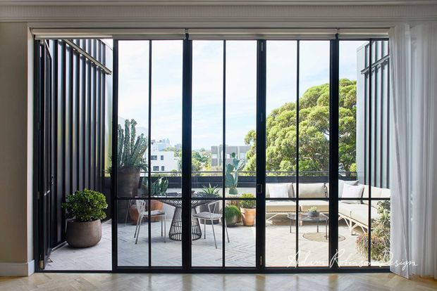 Bright refined interior looking through full-height black-framed glazed doors onto a furnished balcony or terrace. The room has white walls with decorative cornice moulding, light timber flooring, and sheer curtains. Outside is a compact urban terrace with outdoor seating, potted plants, and a city outlook framed by mature trees.