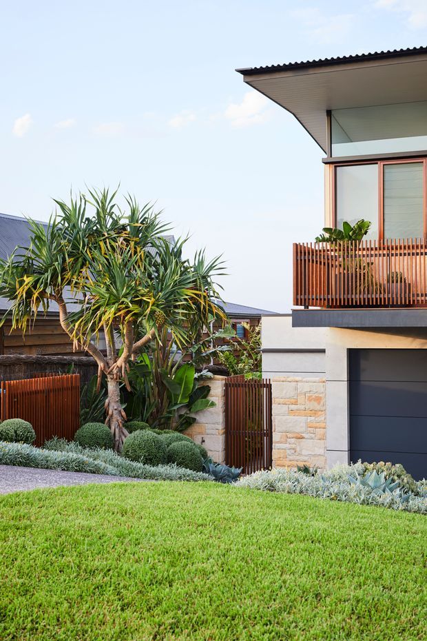Exterior view of a contemporary multi-level residence with a projecting upper-floor roof and balcony, dark-framed glazing, warm vertical timber privacy screening, pale rendered walls, and a light stone-clad lower facade. The property is surrounded by a carefully designed tropical and drought-tolerant landscape with sculptural planting, clipped shrubs, gravel, and a manicured lawn.