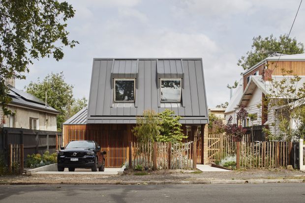 Street-facing view of a compact contemporary multi-storey dwelling in an urban residential setting. The house has a dark grey standing-seam metal-clad upper level with two windows, a lower level screened by vertical timber battens, a small integrated carport or driveway, timber fencing, and neighbouring homes on either side.