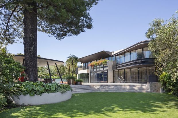 Exterior view of a contemporary multi-level residence set within a landscaped garden. The home features broad glazed walls, projecting roof planes, a curved upper-level roof edge, transparent balcony balustrades, dark framed windows, stone or concrete exterior surfaces, and a wide set of steps leading to the lawn. Mature trees and a planted raised garden bed frame the foreground.