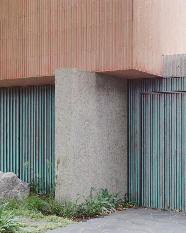 Close-up exterior view of a contemporary building entrance or garage frontage. The facade combines vertically ribbed pink terracotta cladding, dark vertically corrugated metal garage doors, and a rough light-grey rendered concrete pier. Irregular dark stone paving and native-style landscaping with grasses and low plants frame the base, creating a textural, minimalist architectural composition.