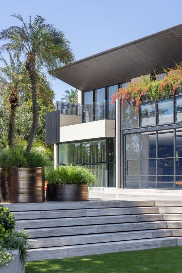 Exterior view of a contemporary multi-level residence with a broad flat projecting roof, extensive dark-framed glazing, cantilevered balcony, and a large glazed garage or entry opening. Wide timber-clad outdoor steps lead up from a manicured lawn to a deck, with large planters and tropical landscaping framing the facade.