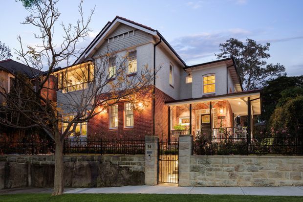 A two-storey residential house photographed from the street at dusk. The facade combines red brickwork on the lower level with light rendered or painted upper walls, a prominent gabled roof, decorative gable detailing, timber-framed windows, and a covered front porch. A low stone boundary wall, black metal fencing, front gate, mature trees, and warm interior lighting complete the suburban streetscape.