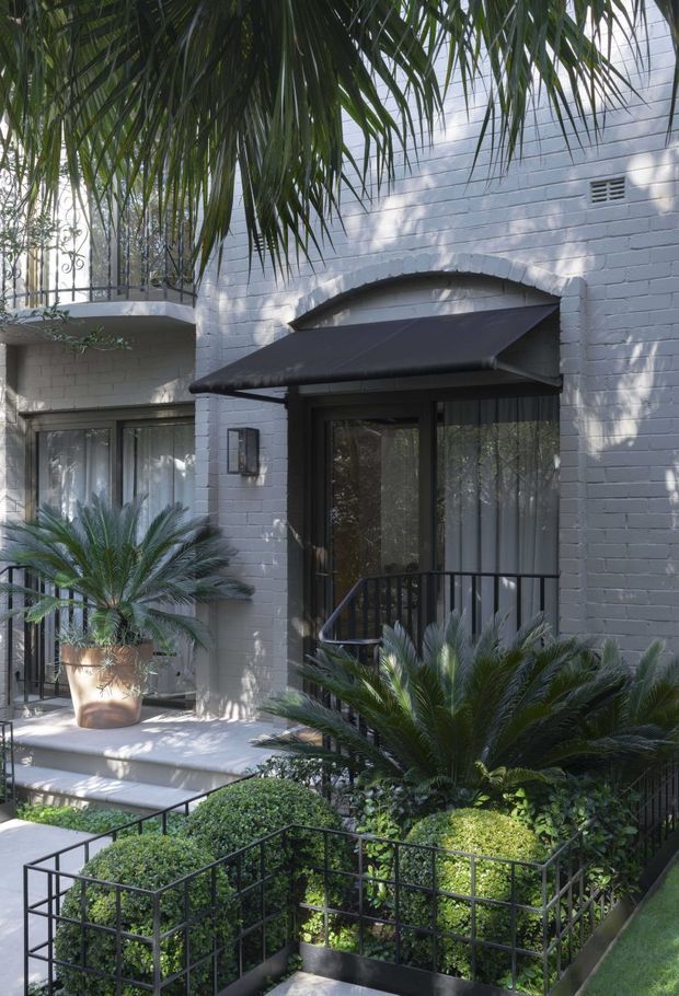 Exterior view of a refined two-storey residential building with a pale painted brick facade, dark framed glazed doors and windows, black metal balustrades, and a dark retractable awning over the entrance. Layered tropical and subtropical planting, including palms in pots and raised garden beds, surrounds the stepped entry courtyard.