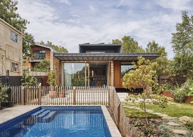 Exterior view of a contemporary multi-level timber-clad house opening onto a landscaped backyard with a swimming pool. The home features extensive glazing, a broad low-pitched roof overhang, timber screens and decking, and a raised terrace with steps leading into the garden. Mature planting and neighbouring residential buildings surround the property.