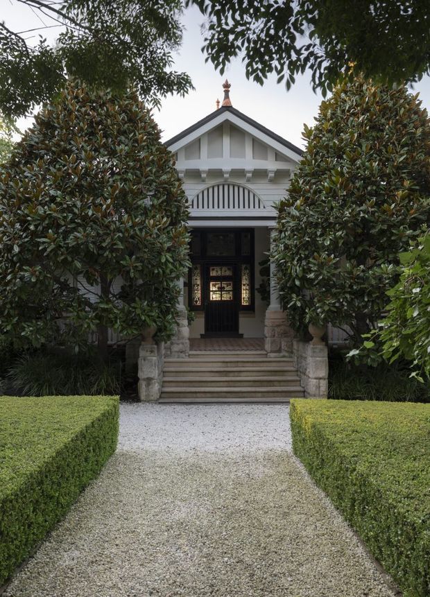 Front exterior view of a formal heritage-style residence, framed by mature hedging and trees. The symmetrical white facade features a steep gabled roof, decorative timber detailing, a small finial, stone-clad porch columns, arched entry ornamentation, a dark timber front door with leadlight glazing, and a stepped gravel path through manicured gardens.