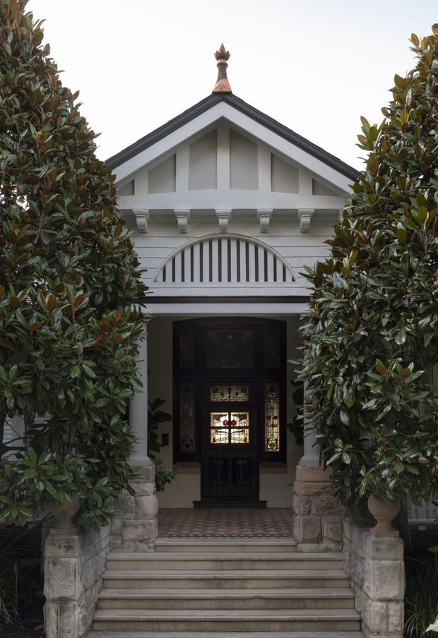 Front elevation of a grand traditional residence, approached by a broad flight of stone steps and framed by mature magnolia trees. The white rendered facade features a steep gabled roof, decorative timber fretwork, classical corbels and columns, a fan-shaped balustrade, and a dark stained-glass entry door. Patterned tiles define the porch floor, while a small ornamental finial crowns the roof.