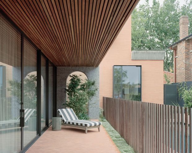 A covered outdoor terrace or balcony beside a contemporary brick residence, featuring full-height dark-framed glazing, a warm timber slatted ceiling, terracotta paving, a striped lounge chair, a textured masonry arch opening, and a vertical timber privacy fence. Mature trees and an adjacent brick building are visible beyond.