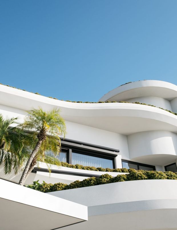 Upward-looking exterior photograph of a bright white multi-level building with sweeping curved balconies, rounded forms, black-framed glazing, planted balcony edges, and a palm tree against a clear blue sky.