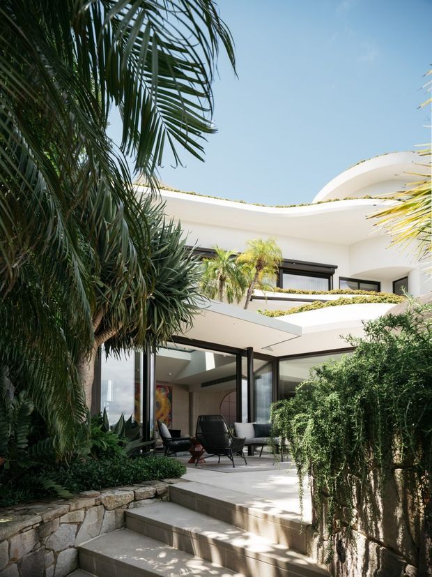 Exterior view of a multi-level contemporary residence surrounded by lush tropical landscaping. The white rendered building has sweeping curved rooflines and projecting terraces, extensive black-framed glazing, planted roof terraces, and an outdoor sitting area accessed by broad stone steps.