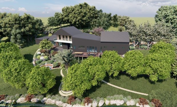Aerial view of a low-rise contemporary residence set within a large landscaped garden. The home has dark pitched roofs, dark rendered or clad walls, extensive glazing, and timber-toned outdoor decking. Curving pedestrian paths, ornamental planting, mature trees, and a rocky garden border surround the house.