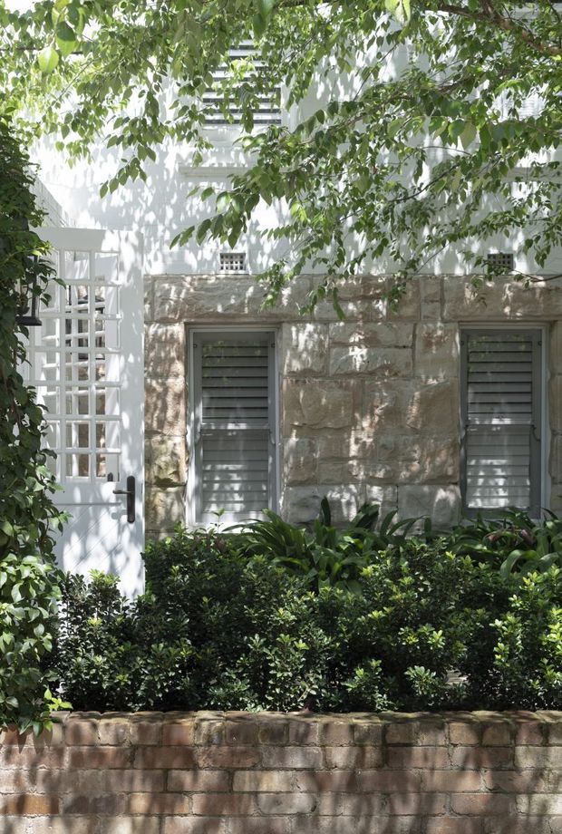 A leafy, shaded view of a refined residential facade. The white rendered upper wall includes a white door with a glazed grid panel and dark hardware, while the lower facade is constructed from large rough stone blocks with grey louvered windows. Dense clipped hedging and layered greenery sit behind a low brick garden wall, creating a private garden frontage.