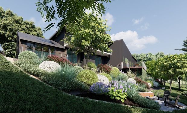 A contemporary multi-level home set into a sloping, heavily landscaped site. The dark-clad building has pitched and gabled roof forms, extensive glazing, timber-toned exterior elements, decks and balconies. Layered ornamental planting, grasses, shrubs and flowering plants dominate the foreground, with an outdoor seating area visible below.