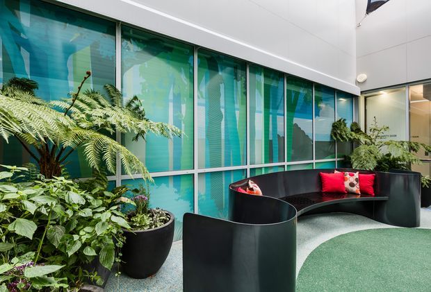 A contemporary indoor courtyard or reception lounge with lush potted tropical planting, curved black built-in seating, colourful cushions, and a large turquoise patterned glazed wall. The space has a bright, playful atmosphere and appears to be part of a commercial interior.