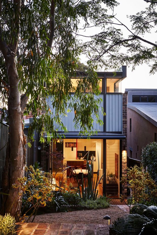 A compact contemporary multi-level residential extension viewed from a landscaped garden courtyard. The house features dark brick walls, corrugated light-grey metal cladding, large glazed openings, and warm interior lighting visible through the ground-floor windows. A brick-paved path, gravel garden, native-style planting, and mature trees frame the facade.