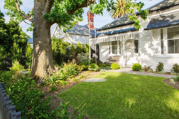 A sunlit heritage-style white weatherboard house viewed from a landscaped front garden, with a broad lawn, mature shade tree, clipped shrubs, garden beds, decorative verandah posts and fretwork, tall windows, and a dark metal pitched roof with a prominent brick chimney.