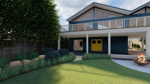 A contemporary elevated residential house viewed from the landscaped front yard. The home features a broad covered lower level supported by slender white columns, dark horizontal cladding, a bright yellow entry door, upper-level balcony glazing and railings, and a distinctive pitched gable roof with exposed triangular framing. The garden includes lawn, layered grasses, shrubs, feature rocks, and a small outdoor fire bowl.