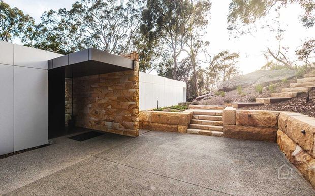 Modern low-profile exterior entrance or courtyard with a dark flat-roofed canopy, white rendered wall panels, and a prominent natural stone feature wall. A textured concrete-paved forecourt leads to broad stone steps and terraced landscaped levels, surrounded by mature trees.
