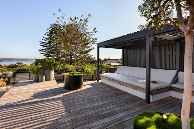 A contemporary outdoor terrace with a weathered timber deck, built-in upholstered bench seating, and a dark metal pergola with a corrugated roof and vertical screening. Large planters and established greenery frame the space, with a distant coastal or waterside outlook visible beyond the terrace.