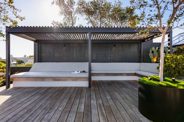 A contemporary outdoor lounge pavilion with built-in white upholstered bench seating, dark vertical screening panels, a slatted pergola roof, and a broad timber deck. Large planters and landscaped greenery frame the relaxed, sunlit space.