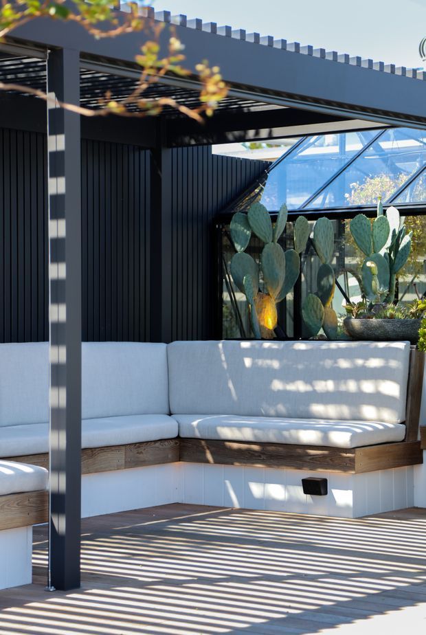 A bright contemporary outdoor lounge beneath a dark pergola, featuring built-in white upholstered bench seating with timber bases, a slatted timber deck, dark vertical wall cladding, and a glazed greenhouse-like enclosure containing large potted prickly pear cacti. Strong linear shadows from the pergola create a high-contrast pattern across the deck and seating.