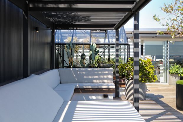 A bright contemporary outdoor lounge beneath a slatted pergola, featuring a large white built-in sectional sofa, timber decking, black metal framing, glass doors, and potted plants. Strong linear shadows create a striped pattern across the seating and deck, with a glazed structure and interior space visible beyond.
