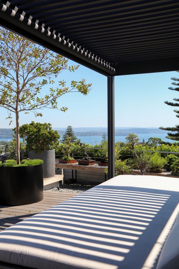 A modern elevated outdoor terrace overlooking a blue bay or waterfront, with a dark slatted pergola casting strong striped shadows across a large pale upholstered daybed or lounge. The terrace includes timber decking, black structural posts and railings, large cylindrical planters, bonsai-style plants, shrubs and a small tree. The scene is sunny, open and contemporary, with distant landscape and water visible beyond the railing.