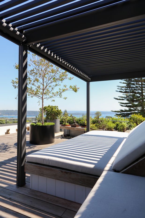 A sunlit outdoor terrace overlooking a body of water, featuring built-in cushioned daybeds beneath a dark louvred pergola. Timber decking, black structural posts, potted plants, and strong striped shadows create a refined coastal outdoor lounge setting.