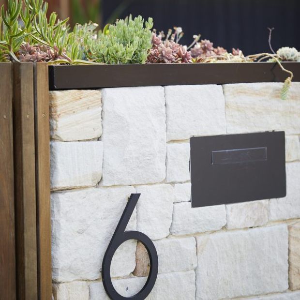 Close-up of a residential exterior mailbox and house number mounted on a pale rough-cut stone wall. A dark rectangular mailbox and matching stylised number sit beneath a dark cap, with a timber side post and succulent planting above.