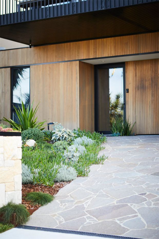 A contemporary residential entry facade with warm vertical timber cladding, dark metal-framed glazing and a projecting upper-level balcony. The entrance is approached by an irregular natural stone-paved path bordered by drought-tolerant landscaped planting and a low stone wall. The design has strong horizontal and vertical lines, with a sheltered recessed doorway and an upper balcony railing visible above.