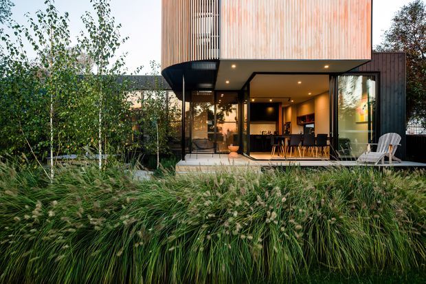 Contemporary two-storey residential extension with a projecting upper volume clad in vertical warm-toned timber battens and a dark lower structure. The open ground floor reveals a kitchen and dining area through extensive glazing, opening onto a paved terrace. Native-style ornamental grasses, slender trees and dense planting frame the building in the foreground.
