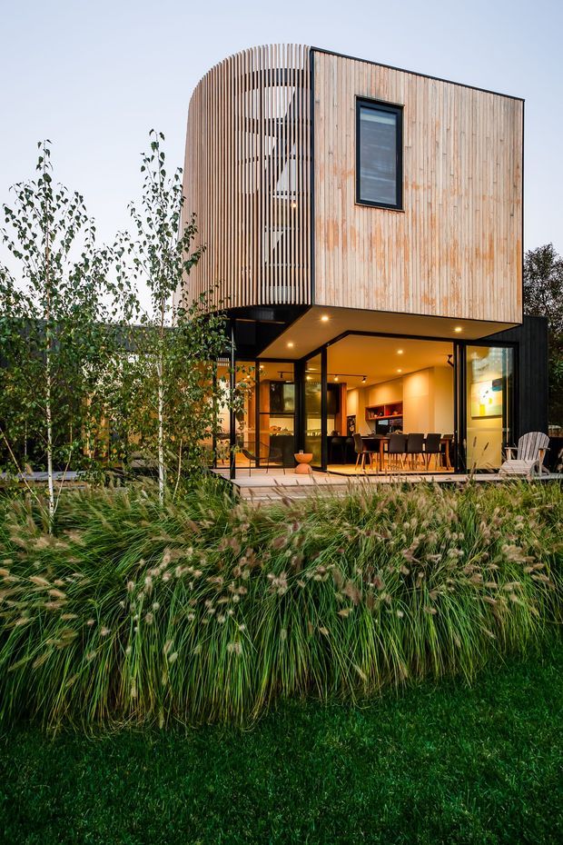 A modern two-storey house viewed from the landscaped garden at dusk. The upper level is clad in vertical light timber boards with a rounded slatted timber screen, while the lower level has dark framing, extensive glazing, and a covered outdoor entertaining area with dining and lounge furnishings. Ornamental grasses and planting occupy the foreground.