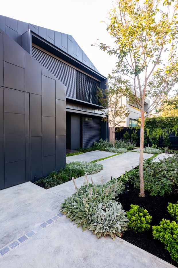 A contemporary residential exterior viewed from a landscaped front garden. The building has dark charcoal panel cladding, a sharply angled roofline, and broad horizontal louvred window screens. Pale concrete paths curve through layered low-maintenance planting, with a mature slender tree and dark boundary fencing.