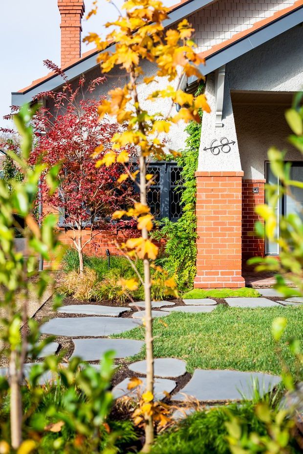 A landscaped front garden and entry to a low-rise residential building. The facade combines red brickwork, textured light render, dark timber or metal-framed glazing, and a steep gabled roof with shingle-like cladding. Irregular large grey stepping stones lead through lawn and layered ornamental planting toward a recessed entrance. Autumn foliage partially frames the view.