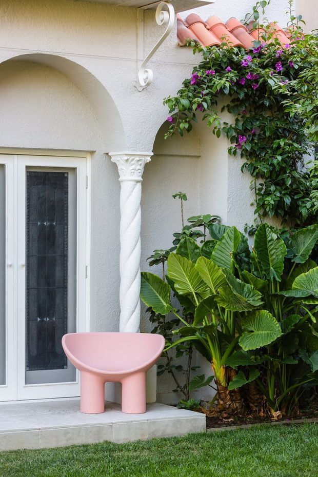 A close-up exterior view of a Mediterranean-influenced stucco facade with a white glazed door, arched openings, decorative twisted column, ornate bracket, terracotta roof tiles, and lush tropical planting. A sculptural pink outdoor stool sits on the tiled entry step.