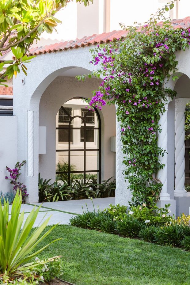Exterior view of a Mediterranean-inspired residence with white stucco walls, rounded arches, twisted columns, and a terracotta tiled roof. A lush flowering vine climbs the front arcade, with manicured lawn and layered garden planting in the foreground.