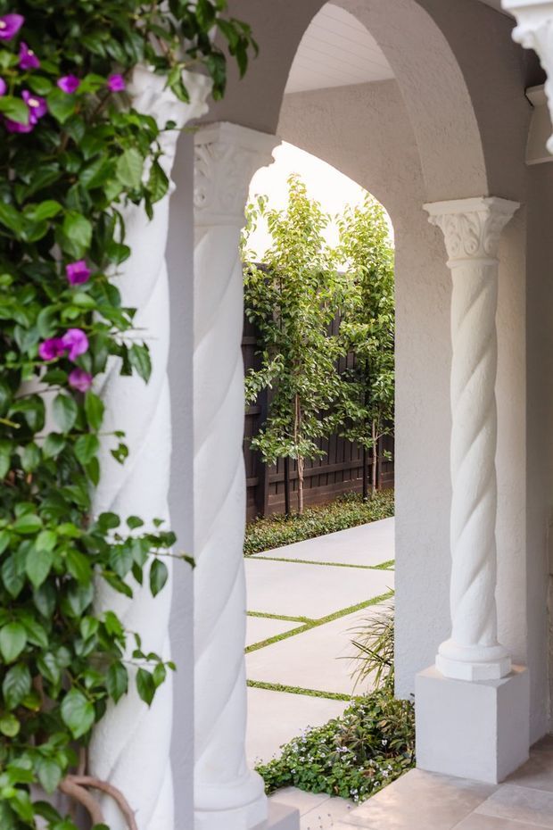 A refined exterior entry arcade with white stucco arched openings and ornate twisted classical columns. A flowering vine climbs the left side, while a landscaped courtyard with pale rectangular paving, clipped planting, and a dark timber fence is visible beyond. The space has a Mediterranean-inspired, elegant character with bright natural light and strong white-and-green contrast.