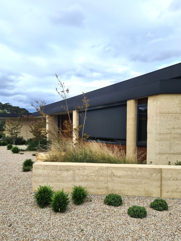 A low-slung contemporary building with a dark charcoal metal-clad roof and facade, pale textured masonry walls, and narrow vertical windows. The building is set within a minimalist gravel landscape featuring sculptural grasses, clipped shrubs, young trees, and low masonry planter walls beneath an overcast sky.