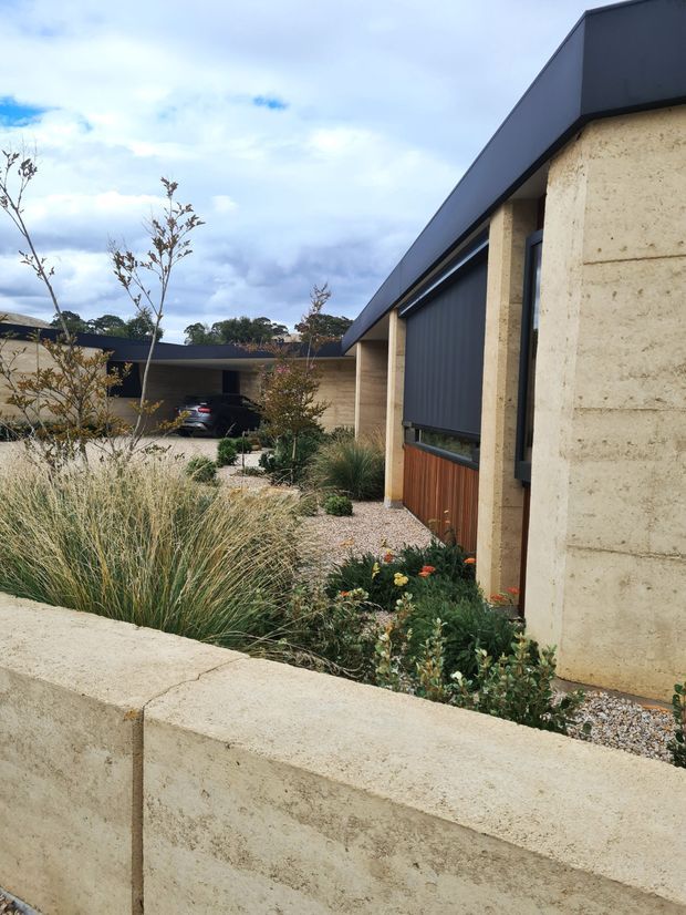 Exterior view of a contemporary single-storey residence with a dark low-pitched roof, broad dark window and door frames, warm timber screening, and textured pale concrete or masonry walls. A landscaped gravel garden with ornamental grasses and planted beds sits in front, with a covered carport visible in the background.