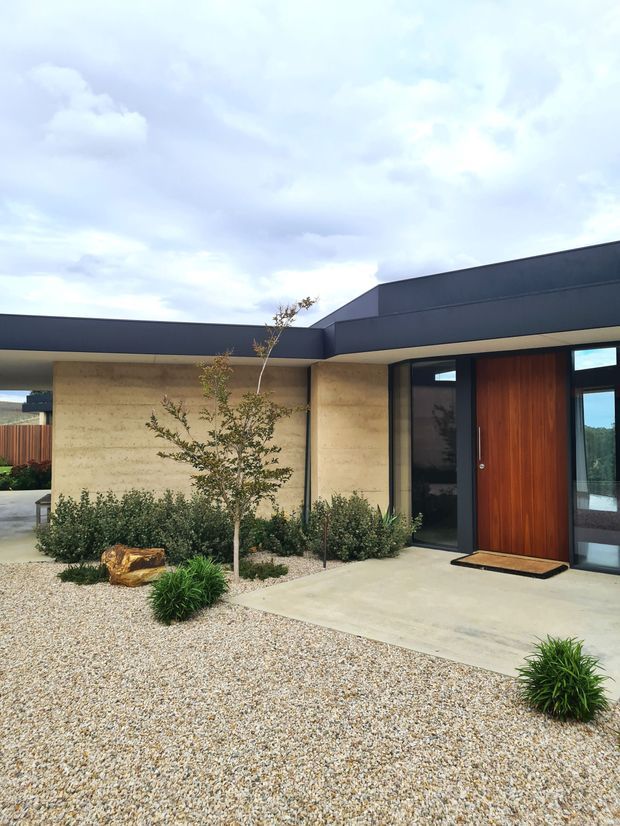 A contemporary single-storey home entrance viewed from the landscaped front courtyard. The facade combines dark flat roof forms, warm timber entry door, large dark-framed glazing, and textured pale concrete or stone walls. The front garden uses pale gravel, clipped shrubs, ornamental grasses, and a small tree, creating a minimalist low-maintenance approach.